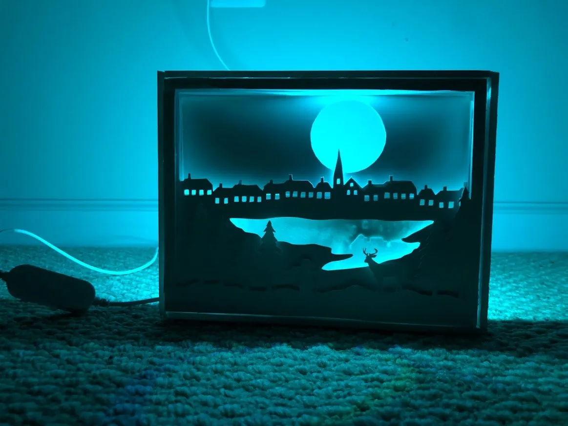 A layered paper lightbox glowing teal: a village skyline and a large moon cut from cardstock, lit by an LED strip.