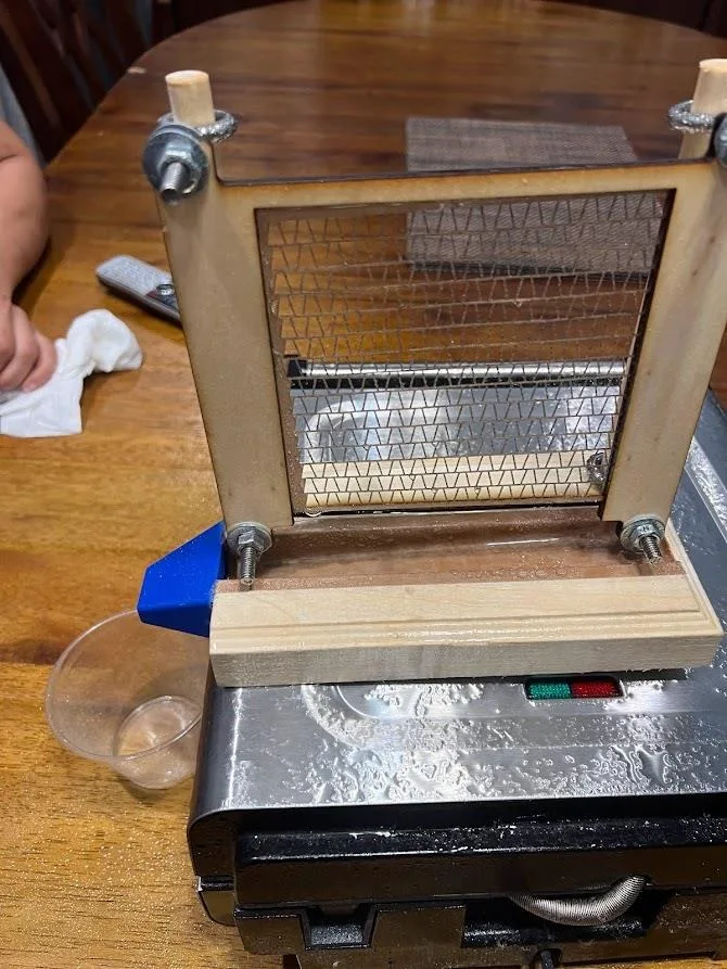 Close view of the rig: a wooden frame holds a square of fine mesh vertically above a base plate with a channel, a blue printed nozzle at its end.