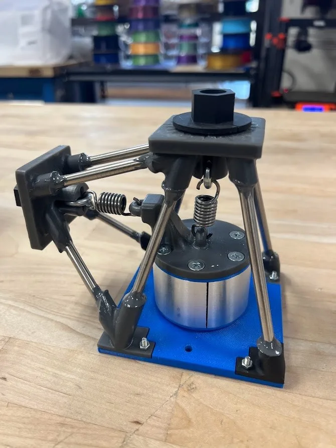 The finished rig: a blue printed base plate holding a cylindrical load cell, with four steel tubes rising to resin-printed corner joints, a vertical spring above the load cell and a horizontal spring at the side, each tensioned by a black knob.