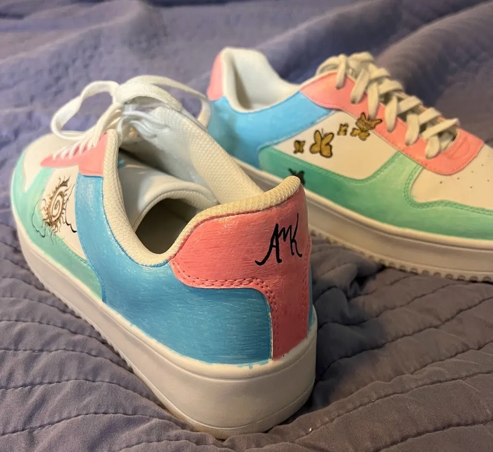 A pair of sneakers hand-painted in pastel pink, blue and green, with a small sun drawn on one heel and butterflies on the other.