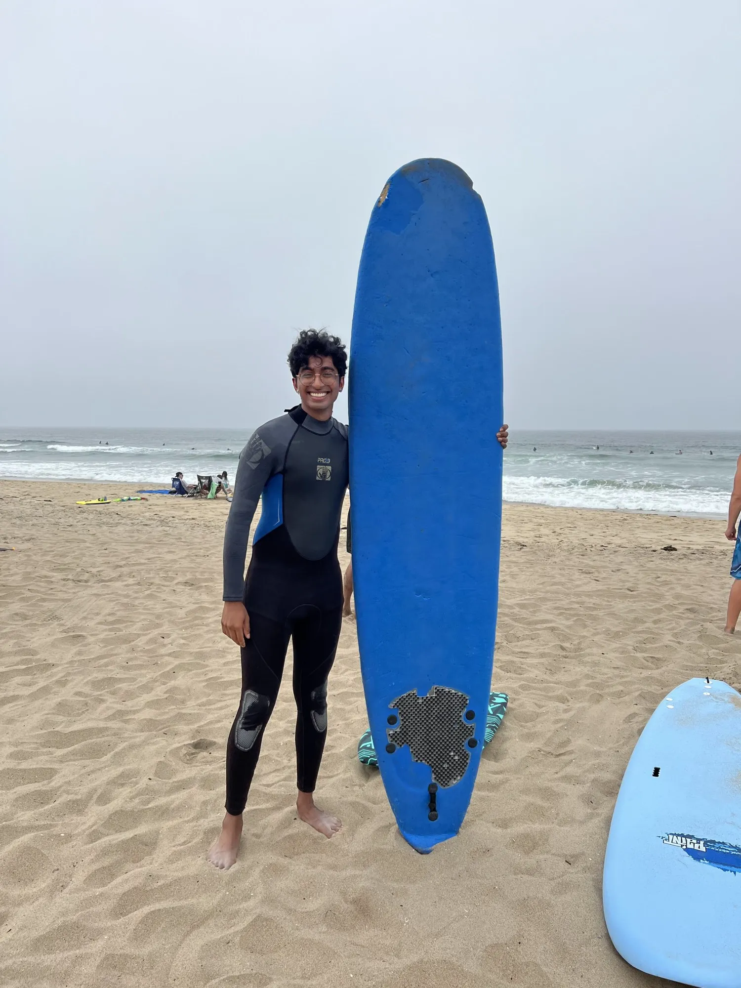 Adithya in a wetsuit on a beach, standing beside a blue longboard taller than him.