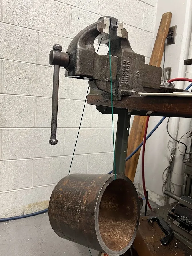 A cylindrical steel weight hanging by a green cord from the loading piece, which is clamped in a bench vise.